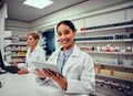 Portrait of smiling young female pharmacist wearing labcoat holding digital tablet with senior colleague working in