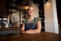 Portrait of smiling young female waitress wearing apron standing leaning on table in cafe Royalty Free Stock Photo