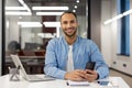 Portrait of a smiling young Hispanic Muslim man sitting in a modern office at a desk, holding a mobile phone and looking Royalty Free Stock Photo