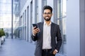 Portrait of a smiling young Indian businessman in a suit standing outside an office building, holding his hand in his Royalty Free Stock Photo