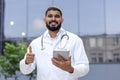 Portrait of a smiling young Indian male doctor, a student standing near a clinical university in a white coat, holding a Royalty Free Stock Photo