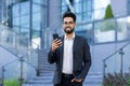 Portrait of a smiling young Indian man in a business suit standing outside a building, holding a phone in his hand, the Royalty Free Stock Photo