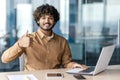 Portrait of a smiling young Indian man sitting at an office desk with a laptop, looking at the camera and showing a Royalty Free Stock Photo