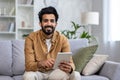 Portrait of a smiling young Indian man sitting on a sofa, holding a tablet and looking at the camera Royalty Free Stock Photo
