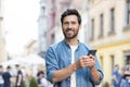 Portrait of a smiling young man standing on a city street, looking at the camera and holding a mobile phone Royalty Free Stock Photo