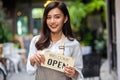 Portrait of a smiling young woman in apron holding open sign board while standing at the cafe. The coffee shop is raising the sign