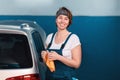 Portrait of smiling young woman in blue coveralls. Car and workshop at the background. Work at auto repairshop and