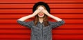 Portrait of smiling young woman covering her eyes with her hands wearing a black round hat, white striped shirt on red wall Royalty Free Stock Photo