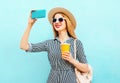 Portrait smiling young woman with cup of juice taking a selfie by smartphone wearing a summer straw hat and backpack on a blue Royalty Free Stock Photo