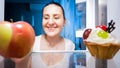 Closeup portrait of smiling young woman doubting between eating apple or cake at night Royalty Free Stock Photo