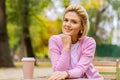 Portrait of smiling young woman drinking coffee at the terrace cafeteria in the park. Blonde female in the city street drinking a