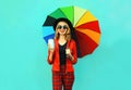 Portrait smiling young woman holding coffee cup and colorful umbrella in red jacket, black hat on blue wall Royalty Free Stock Photo