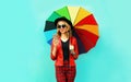 Portrait smiling young woman holding phone with colorful umbrella in red jacket, black hat on blue wall Royalty Free Stock Photo