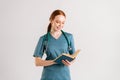 Portrait of smiling young woman intern in green uniform with stethoscope reading medical book standing on white isolated Royalty Free Stock Photo