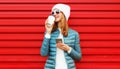 Portrait smiling young woman with phone and cup of coffee wearing a jacket and white hat on red background Royalty Free Stock Photo
