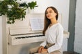 Portrait of smiling young woman pianist looking at camera sitting at classical white piano in classroom. Royalty Free Stock Photo