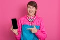 Portrait of smiling young woman pointing with finger on blank smartphone screen isolated over pink background, girl with ponytail Royalty Free Stock Photo