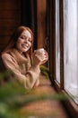 Portrait of smiling young woman standing near window with cup hot coffee and looking at camera. Royalty Free Stock Photo