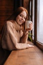 Portrait of smiling young woman standing near window with cup hot coffee and looking at camera. Royalty Free Stock Photo