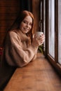 Portrait of smiling young woman standing near window with cup hot coffee and looking at camera. Royalty Free Stock Photo