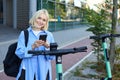 Portrait of smiling young woman unlocks scooter on street, using mobile phone to rent it with smartphone application