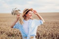 Portrait of smiling young woman walking among wheat in summer field wearing straw hat holding bundle of ripe wheat