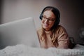 Portrait of smiling young woman wearing headphones listening to music on her laptop at home while lying on bed working Royalty Free Stock Photo