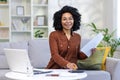 Portrait of a smiling young woman working remotely. Sitting on the sofa in front of the laptop, holding documents and Royalty Free Stock Photo