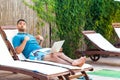 Portrait of smug handsome bearded young adult man in blue t-shirt and shorts lying on cozy sunbed with laptop on poolside and Royalty Free Stock Photo