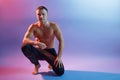 Portrait of sportsman squatting against neon background, wearing black trousers, posing topless and barefoot, showing his perfect Royalty Free Stock Photo