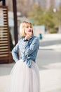 A portrait of a standing dreamy blonde girl with bright pink lips hoding her hand near chin wearing denim shirt, grey