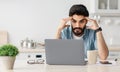 Portrait of stressed pensive arab man sitting at desk with laptop in kitchen, depressed freelancer touching temples