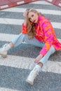 Portrait of striking blonde in bright blouse in coral orange tones, jeans and high shoes, sitting on asphalt in city.