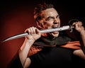 Portrait of strong courage fervor man warrior in cape holding sabre, sword in his mouth, ready to fight over red Royalty Free Stock Photo