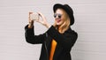 Portrait stylish cheerful laughing young woman taking selfie by smartphone wearing a black coat, round hat on gray background Royalty Free Stock Photo