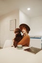 Portrait of a stylish girl in a hat sits in a cozy cafe, petting a dog and drinking coffee. Lady with a pet in her arms spends Royalty Free Stock Photo