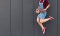 Portrait of a stylish girl in a jeans sarafan and red sneakers that jumps against the background of a dark wall