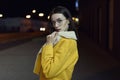 Stylish girl posing in yellow jacket and glasses, lit by city centre lights by night. Womenswear fashion. Bokeh background