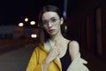 Portrait of stylish girl, posing in yellow jacket, glasses, lit by city centre lights. Womenswear fashion