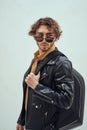 Portrait of a stylish guy with curly hair posing in the bright studio wearing sunglasses, leather coat and backpack Royalty Free Stock Photo