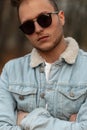 Portrait of a stylish handsome young hipster man in sunglasses in a vintage blue denim jacket with a trendy hairstyle outdoors in Royalty Free Stock Photo