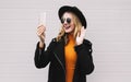 Portrait stylish happy laughing young woman having video call or taking selfie by smartphone wearing a black coat, round hat on Royalty Free Stock Photo