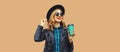 Portrait of stylish happy smiling young woman holds cup of coffee wearing round hat and rock style black leather jacket posing on Royalty Free Stock Photo