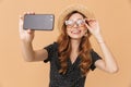 Portrait of stylish joyful woman wearing straw hat and sunglasses smiling and taking selfie photo on mobile phone, isolated over