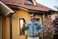 Portrait of stylish man in glasses and jeans jacket, with mobile phone posed against his house