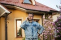 Portrait of stylish man in glasses and jeans jacket, with mobile phone posed against his house