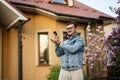 Portrait of stylish man in glasses and jeans jacket, with mobile phone posed against his house