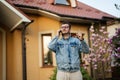 Portrait of stylish man in glasses and jeans jacket, with mobile phone posed against his house and show finger up
