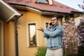 Portrait of stylish man in glasses and jeans jacket, with mobile phone posed against his house and show hand gesture left side