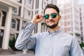 Portrait of stylish man in sunglasses on street in city. He wears blue shirt, beard, smiling to camera.